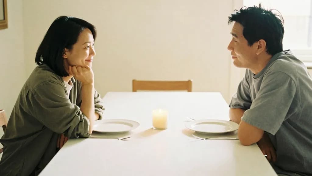 An East Asian woman in her mid thirties with dark hair in a short bob is sitting at one end of a long dining table with one other person at the far end, the…
