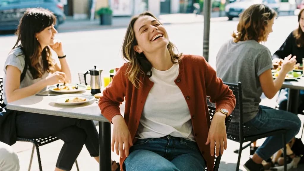 A white woman in her late twenties with shoulder-length dark blonde hair is at a crowded weekend brunch table on a sunlit patio with two friends, plates and…