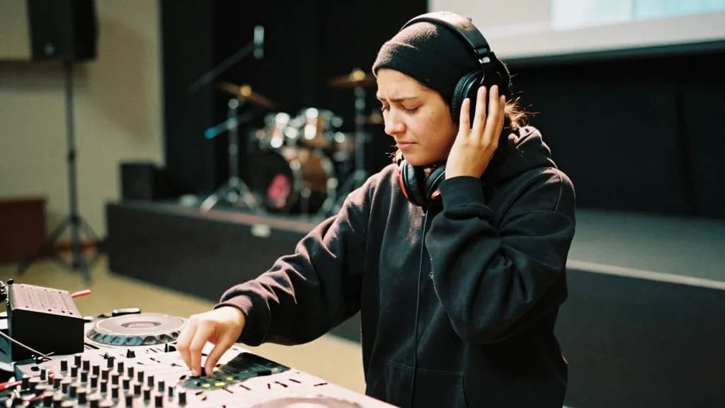 A Latina woman in her late twenties with dark hair under a beanie is mid soundcheck with one hand on a fader and the other pressed to one ear of her headphon…