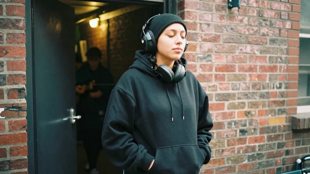 A Latina woman in her late twenties with dark hair under a beanie is standing outside the venue's fire exit in an alley during the support act, the door prop…