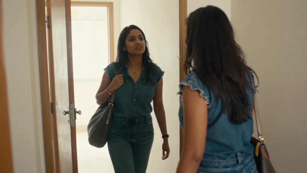A South Asian woman in her late twenties with long dark hair is standing in the hallway fully dressed checking herself in the mirror before leaving, a bag ov…