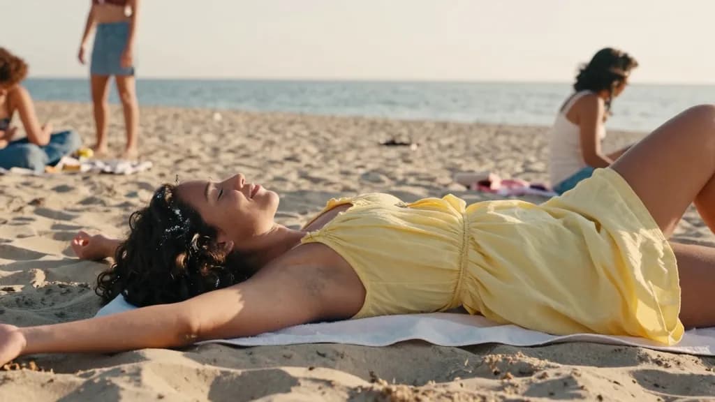 A Latina woman in her late twenties with dark curly hair loose is lying on a towel on the sand in the late afternoon with her eyes shut and salt-dried hair,…