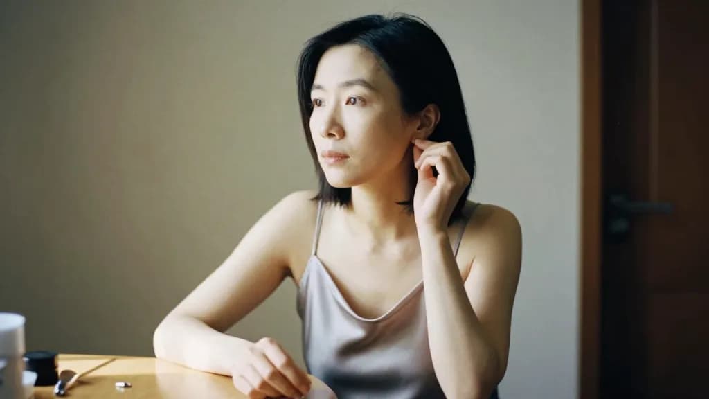 An East Asian woman in her thirties with a sleek shoulder-length bob is sitting at the dresser mid preparation with one earring in, the other still on the su…