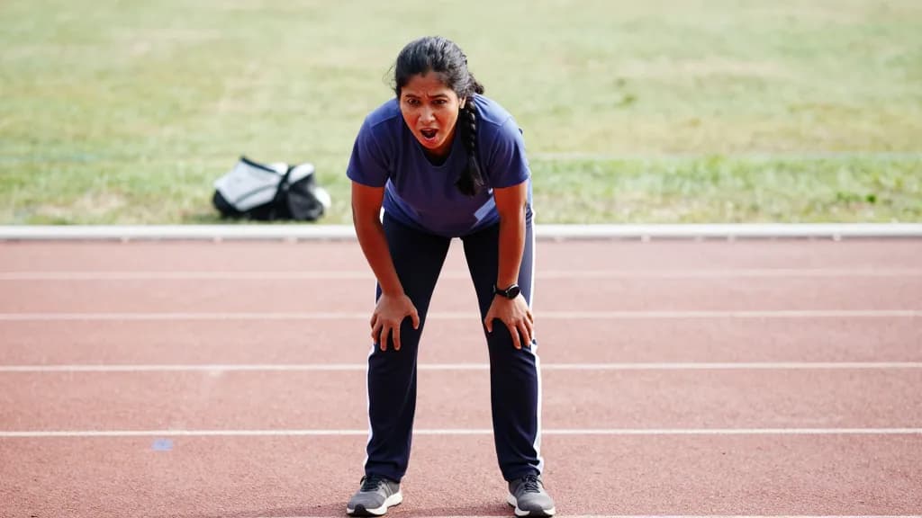 A South Asian woman in her forties with hair braided back is doubled over at the side of a track with her hands on her knees after a hard effort, an empty la…