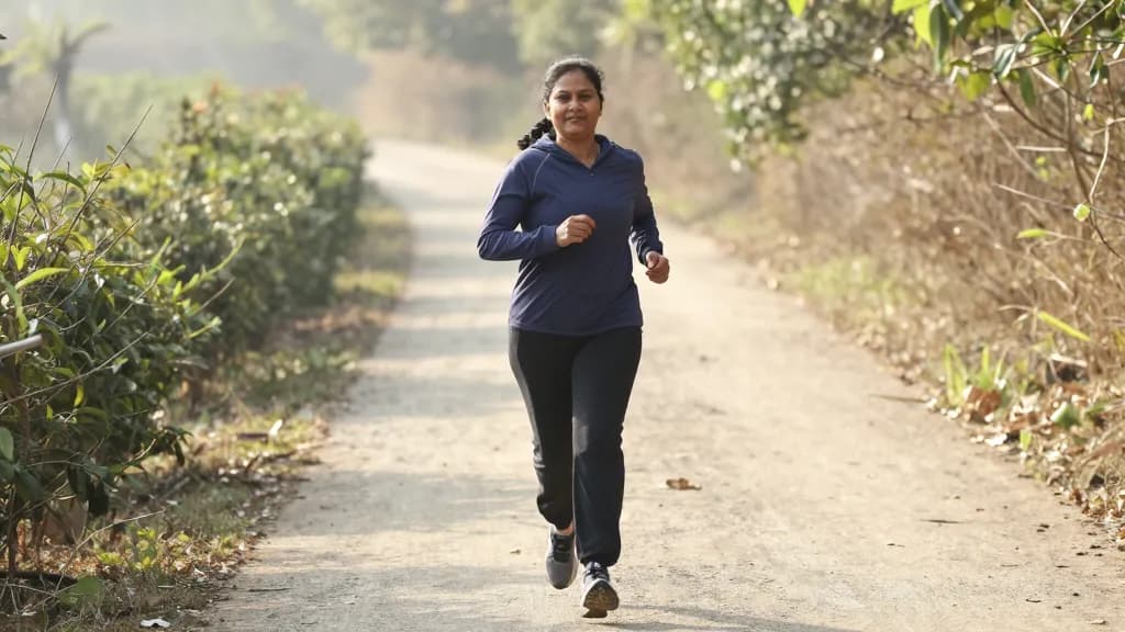 A South Asian woman in her forties with hair braided back is running steadily along the trail in good light with an easy rhythm, hedges either side and the p…
