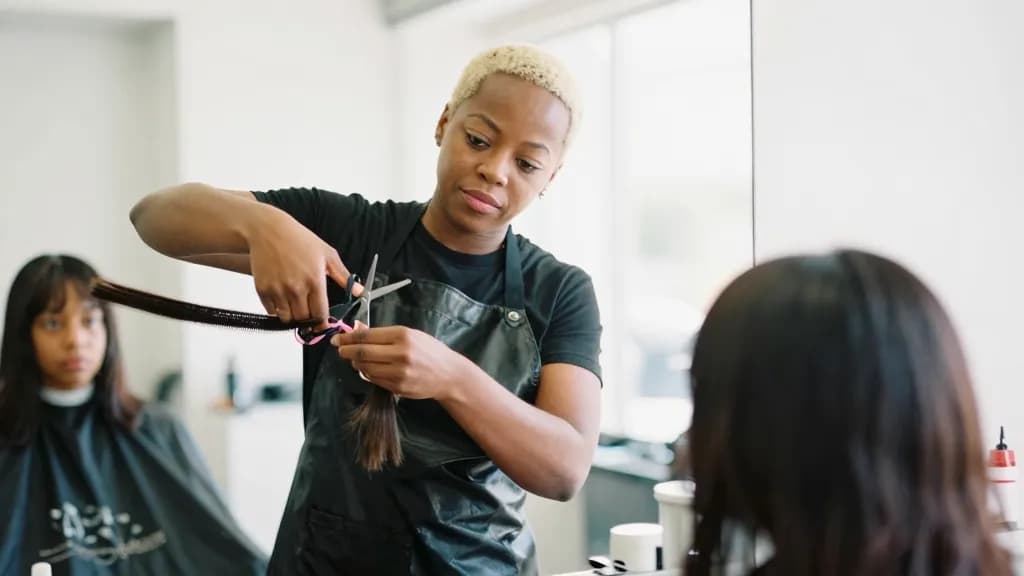 A Black woman in her thirties with short bleached hair is trimming a fraction from a client's ends with scissors, the cut barely different and the client wat…