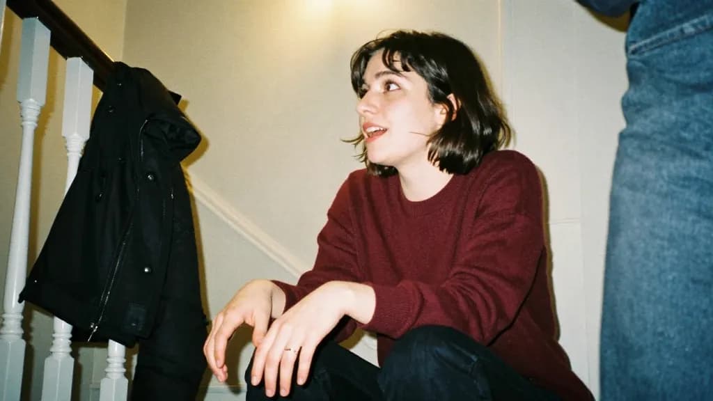 A white woman in her early thirties with dark bobbed hair is sitting on a stairway at the party mid conversation with someone beside her, coats piled on the…