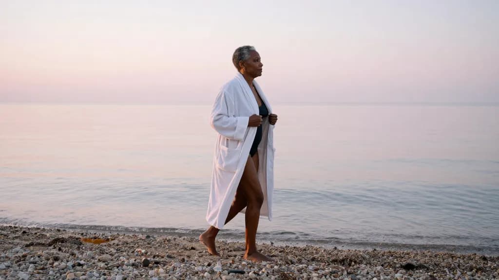 A Black woman in her fifties with short grey-flecked hair is standing alone on a shingle beach at first light with the robe still on and the sea flat in fron…