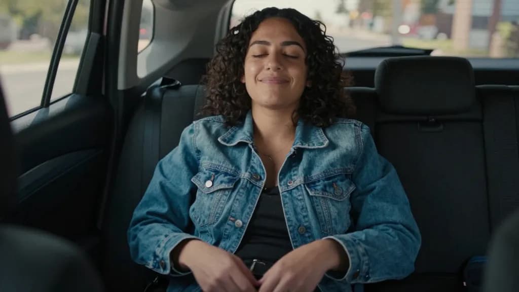 A Latina woman in her late twenties with dark curly hair is sitting in the car with her eyes closed and a small smile, hands loose in her lap, the song clear…