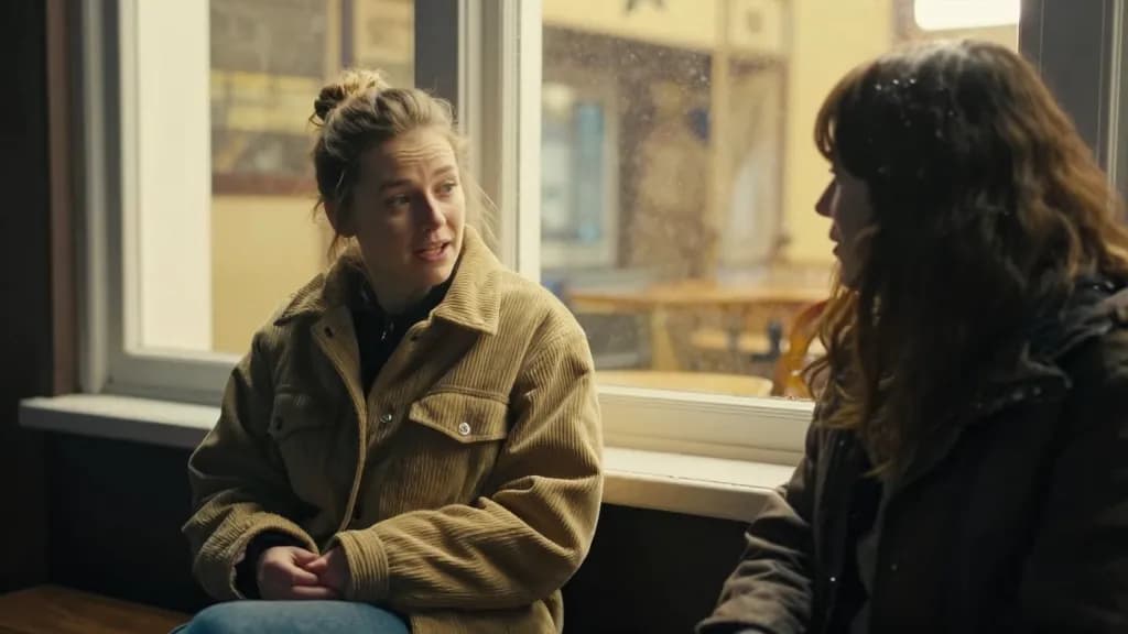 A white woman in her thirties with hair in a messy bun is sitting on a low wall outside the pub in the cold with a friend, both of them talking properly, the…