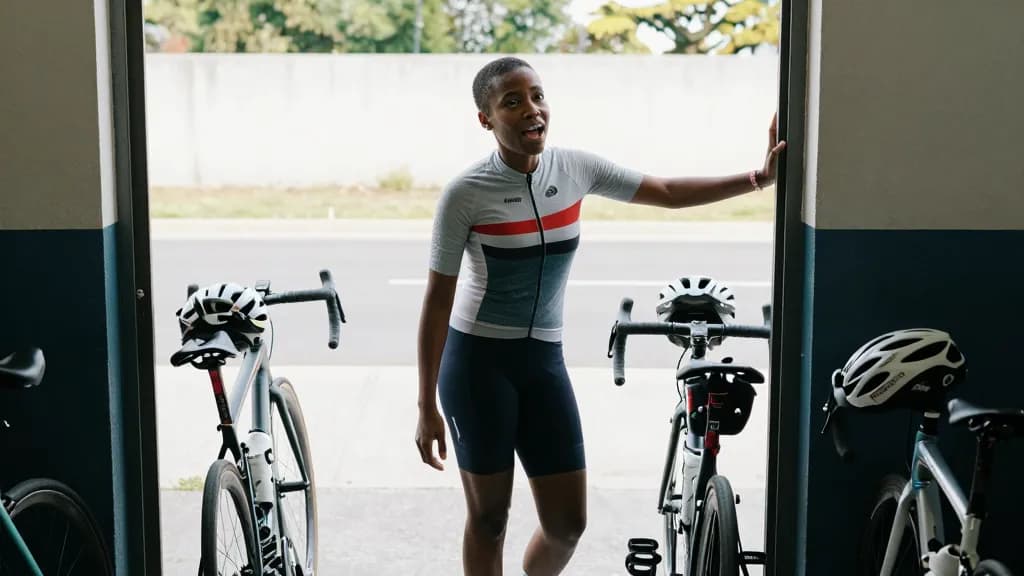 A Black woman in her early thirties with short natural hair is standing in the depot doorway briefing three riders before the shift, bikes leaning and helmet…