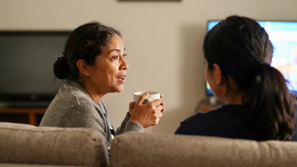 A Latina woman in her forties with dark hair in a low bun is sitting on a sofa beside her partner in the evening with a mug, the television off and both of t…