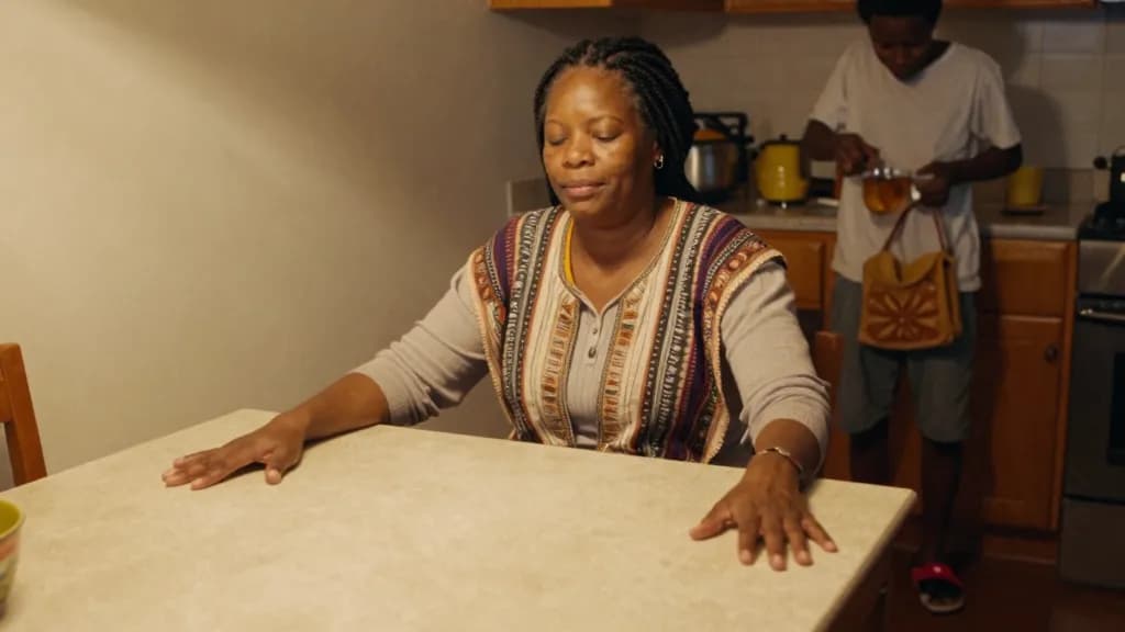 A Black woman in her forties with braids gathered up is sitting at her own kitchen table at home while her adult son makes tea behind her, her shoes off and…