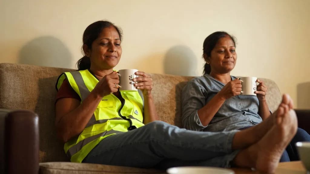 A South Asian woman in her forties with dark hair tied back is sitting on a sofa at home with her shoes off and her feet up, a mug in both hands and her part…