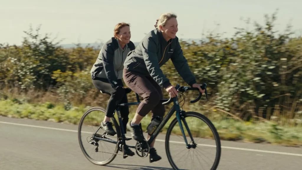 A white woman in her forties with sandy hair in a plait is riding the tandem on a flat road with her partner in front, both of them pedalling in perfect time…