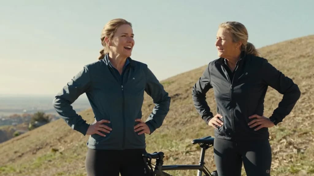 A white woman in her forties with sandy hair in a plait is standing beside the stopped tandem halfway up the hill with her partner, both of them out of breat…