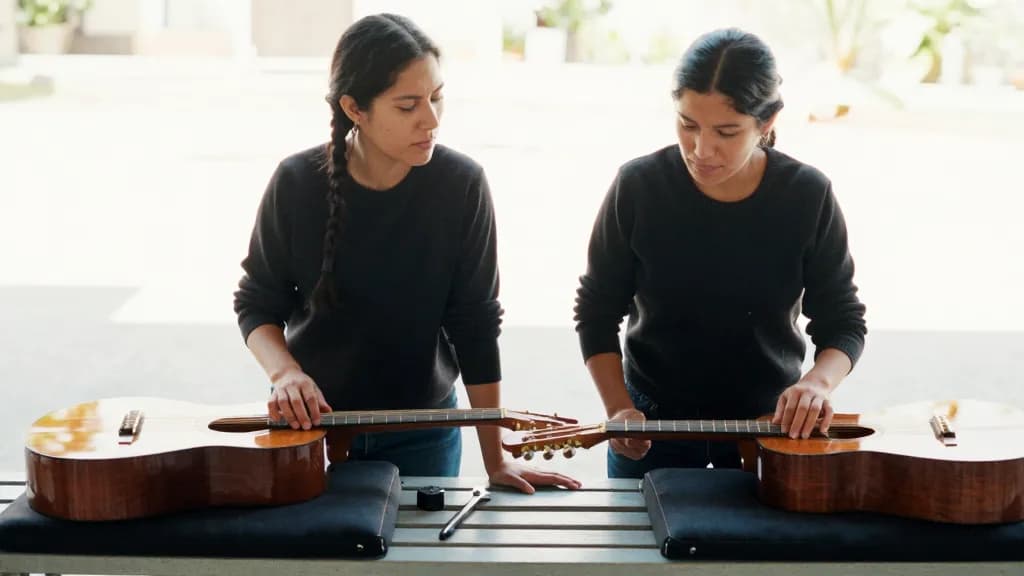 A Latina woman in her thirties with dark hair in a plait is standing at the bench with two similar guitars laid side by side on padded rests, tools between them