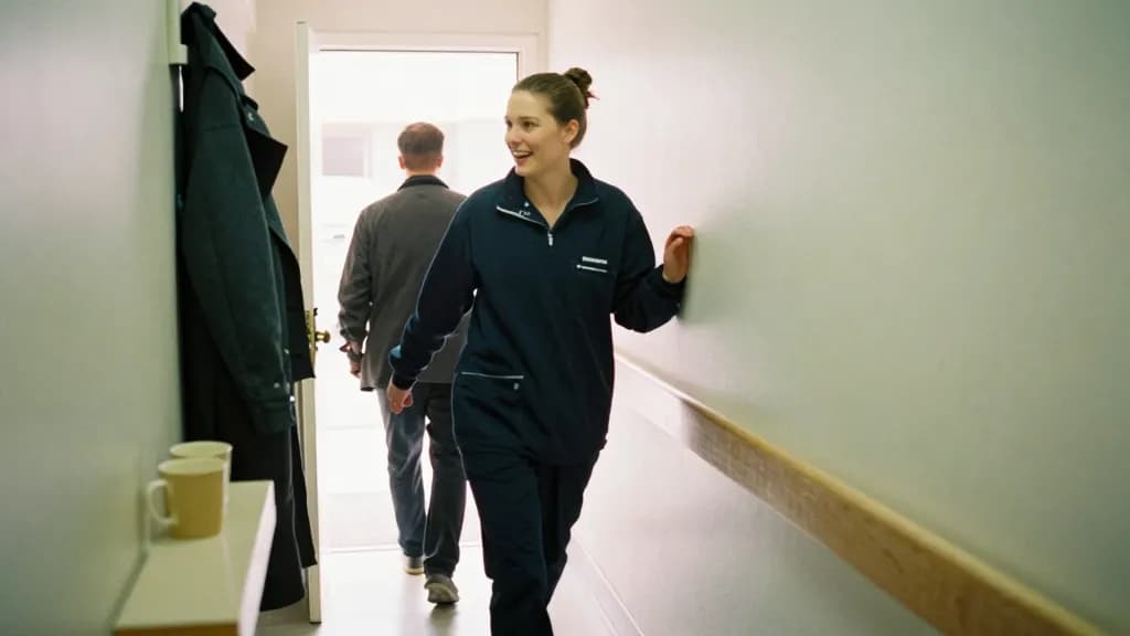 A white woman in her early thirties with hair scraped into a bun is passing her partner in the narrow hallway, one coming in and one going out, coats crowdin…