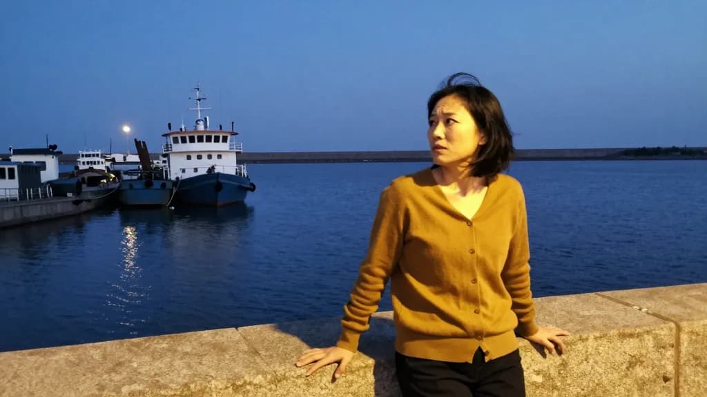 An East Asian woman in her early thirties with a dark bob is standing on a harbour wall at dusk watching a boat that has drifted well off its mooring line, o…
