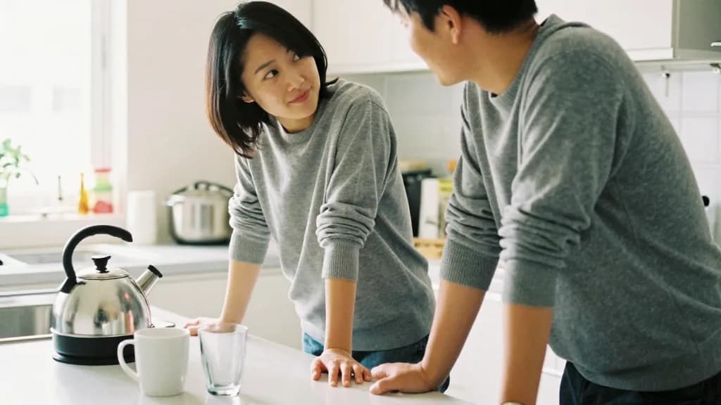 An East Asian woman in her late twenties with a dark bob is standing at a kitchen counter with her fiancé beside her, both of them looking at the same thing…