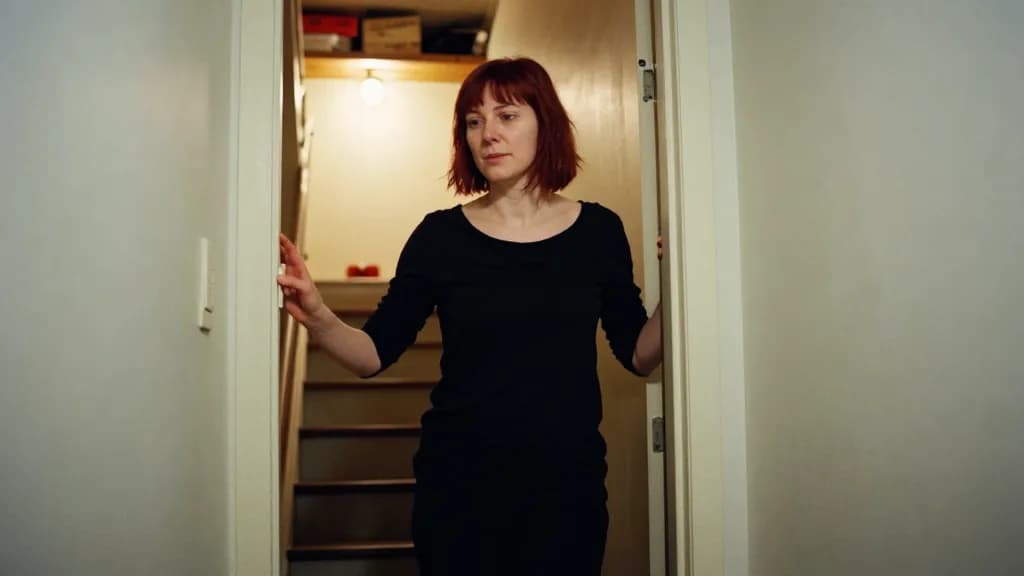 A white woman in her early forties with dark red hair cut short is standing at the top of a dim basement staircase with the light off below her, a hand on th…