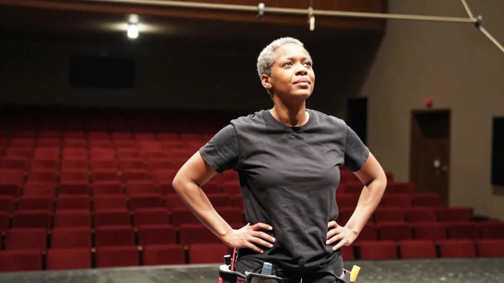 A Black woman in her early fifties with grey-flecked hair cropped short is standing in the middle of an empty theatre stage under a single working light, row…