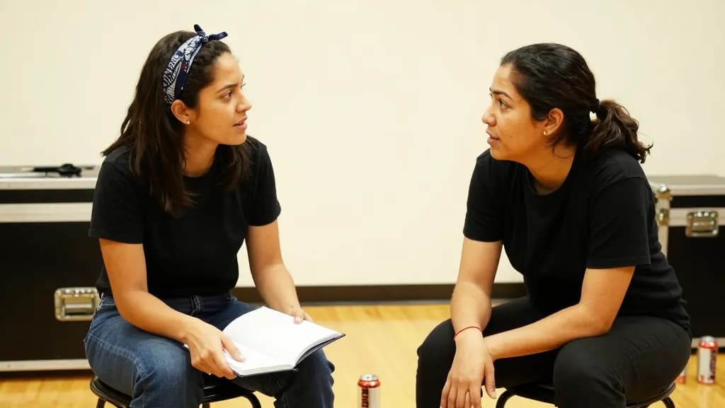 A Latina woman in her early thirties with dark hair pushed back with a bandana is sitting on a flight case in the rehearsal room across from her bandmate on…
