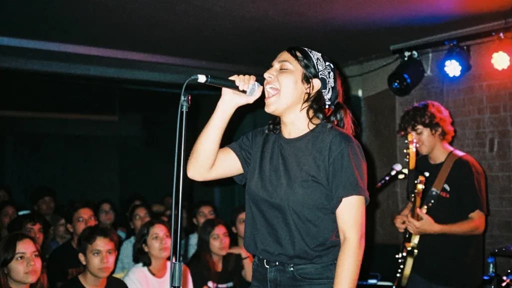 A Latina woman in her early thirties with dark hair pushed back with a bandana is on a small club stage mid set with her bandmate beside her, stage lights wa…