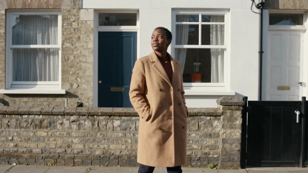 A Black woman in her mid thirties with short natural hair is standing on the pavement outside a modest terraced house she grew up in, a low wall and a gate i…