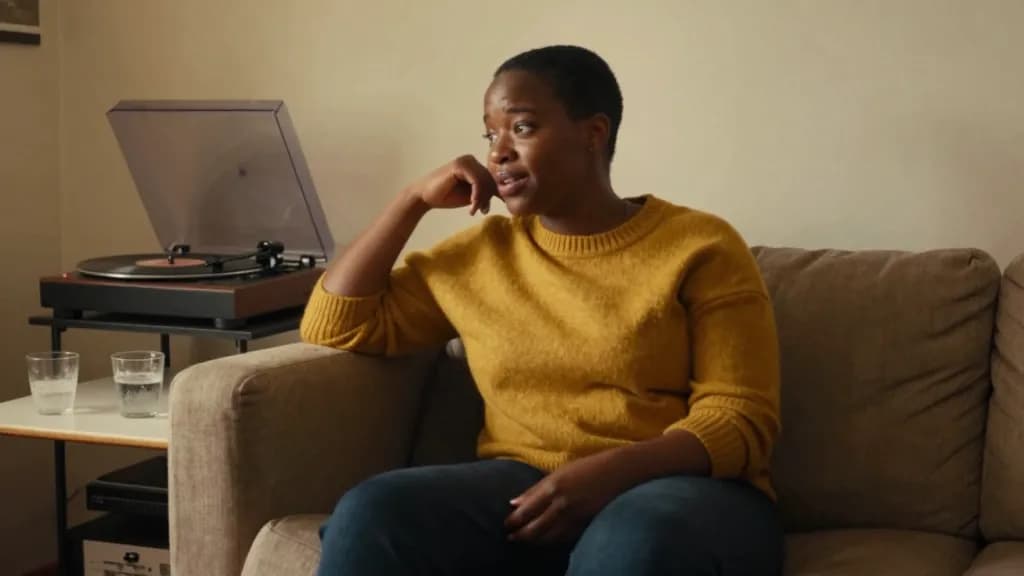 A Black woman in her mid thirties with short natural hair is sitting on the arm of a sofa in a warm front room mid conversation with someone off frame, a rec…