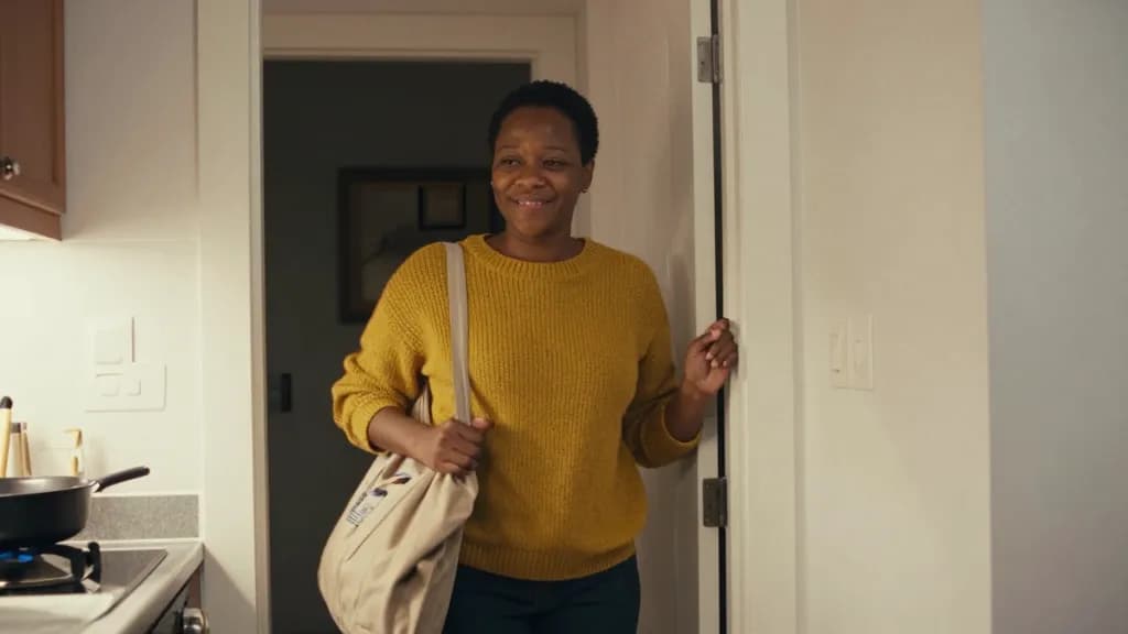 A Black woman in her mid thirties with short natural hair is standing in a kitchen doorway on a weekday evening watching someone cook out of frame, a bag sti…