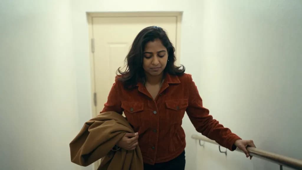 A South Asian woman in her early thirties with dark hair cut to the shoulder is walking out of a party into a stairwell alone, the noise still coming through…