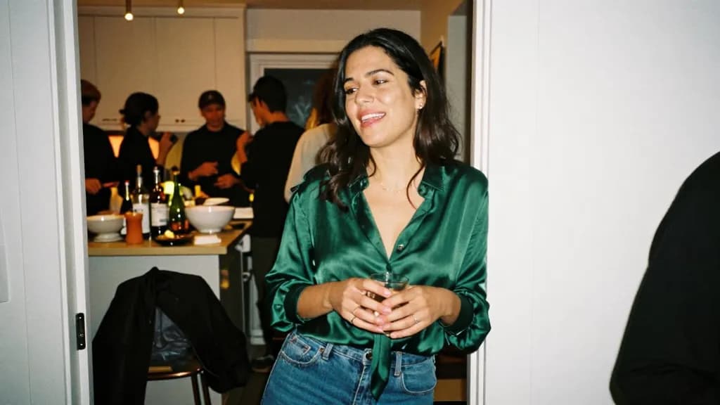 A Latina woman in her early thirties with dark hair worn loose is at a crowded house party in a kitchen doorway, people packed around a counter of bottles an…