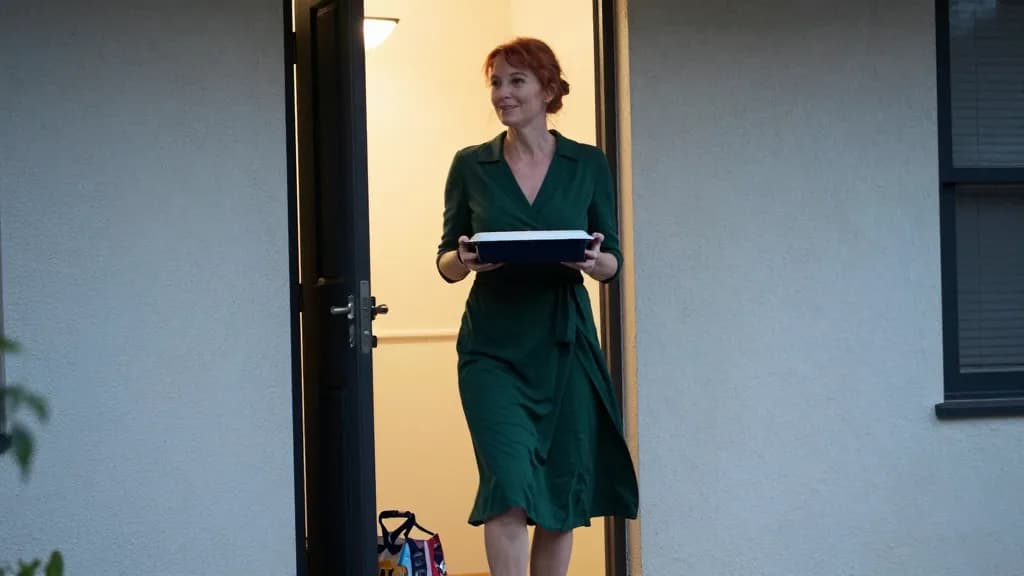 A white woman in her forties with copper hair in a loose knot is standing on a doorstep holding a covered casserole dish in both hands, a carrier bag at her…