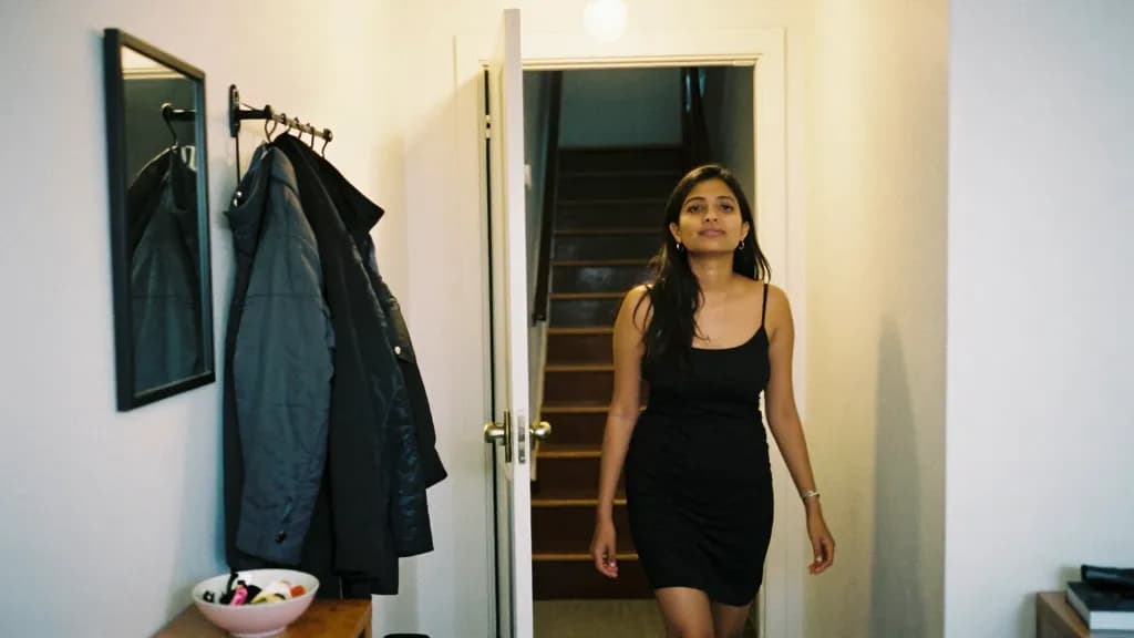 A South Asian woman in her mid twenties with long dark hair worn loose is standing in a narrow hallway by the front door of an apartment, a coat rack loaded…