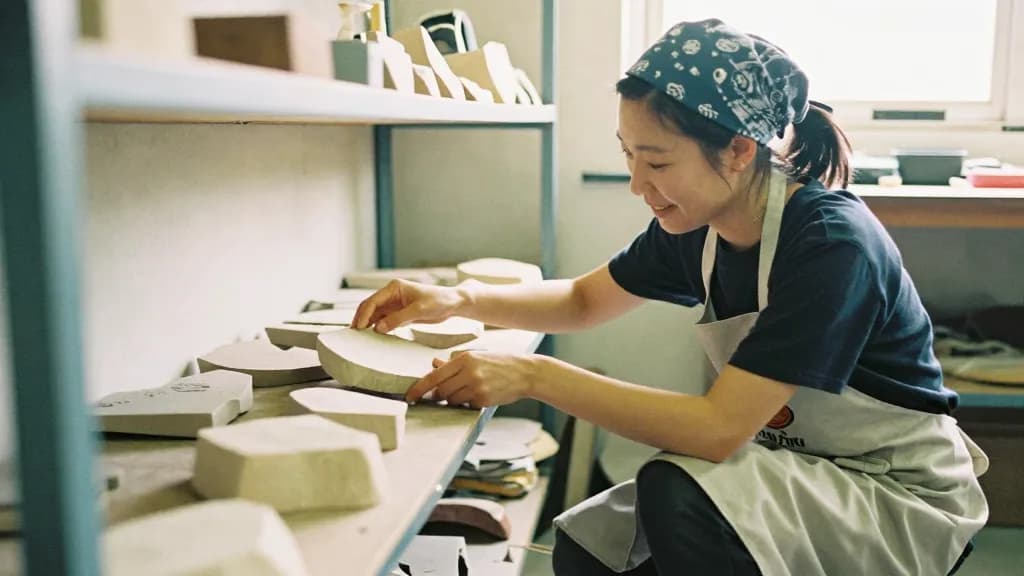 An East Asian woman in her twenties with hair tied back under a bandana is placing a finished uneven piece into the annealer among better ones, the shop quie…