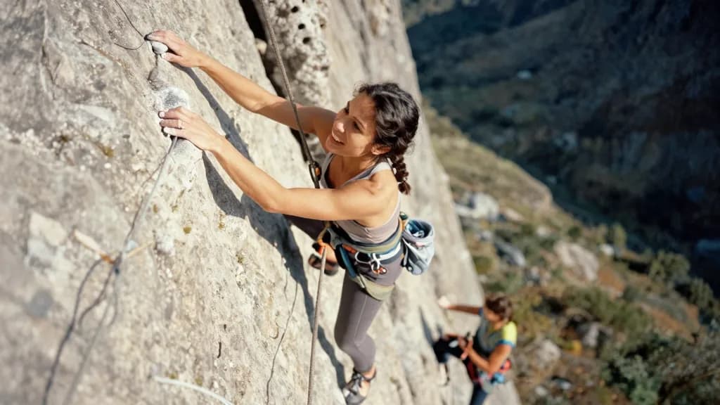 A Latina woman in her late twenties with dark hair in a French plait is climbing a steep section of rock with the rope running below her, her partner belayin…
